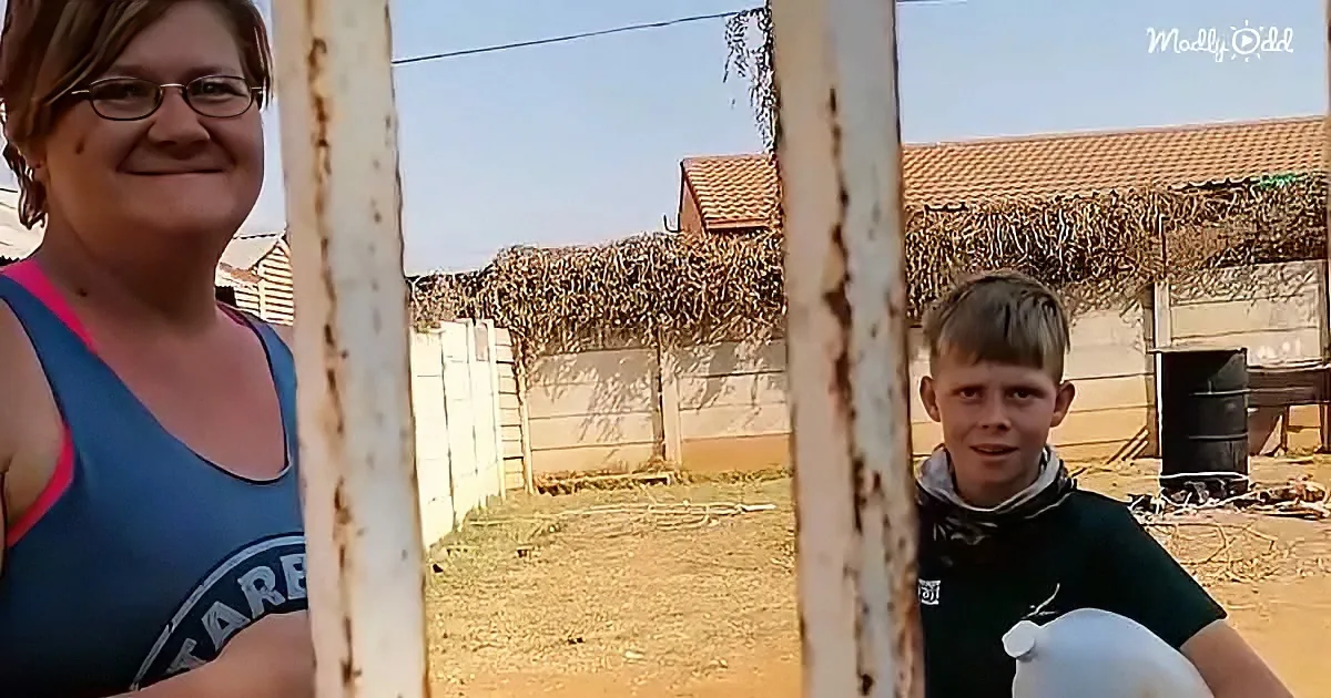 A woman and a boy smile behind a metal gate in a rural setting with thatched roofs and a clear sky.
