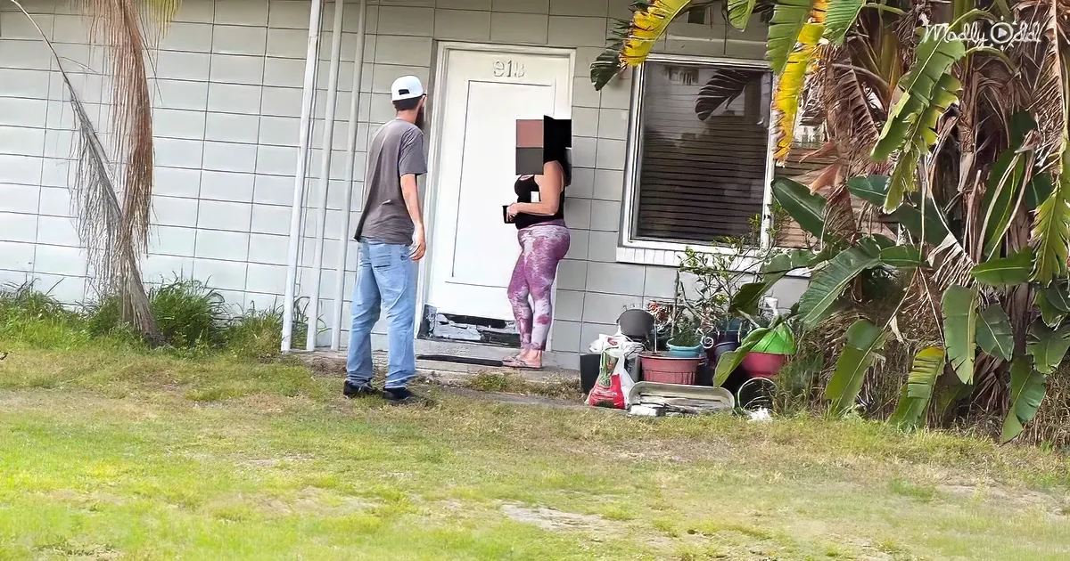A man and woman stand outside a house with a white door, near potted plants and grass.