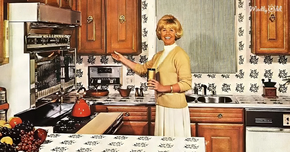 A woman in a vintage kitchen smiles, holding a drink, with retro appliances and floral wallpaper.