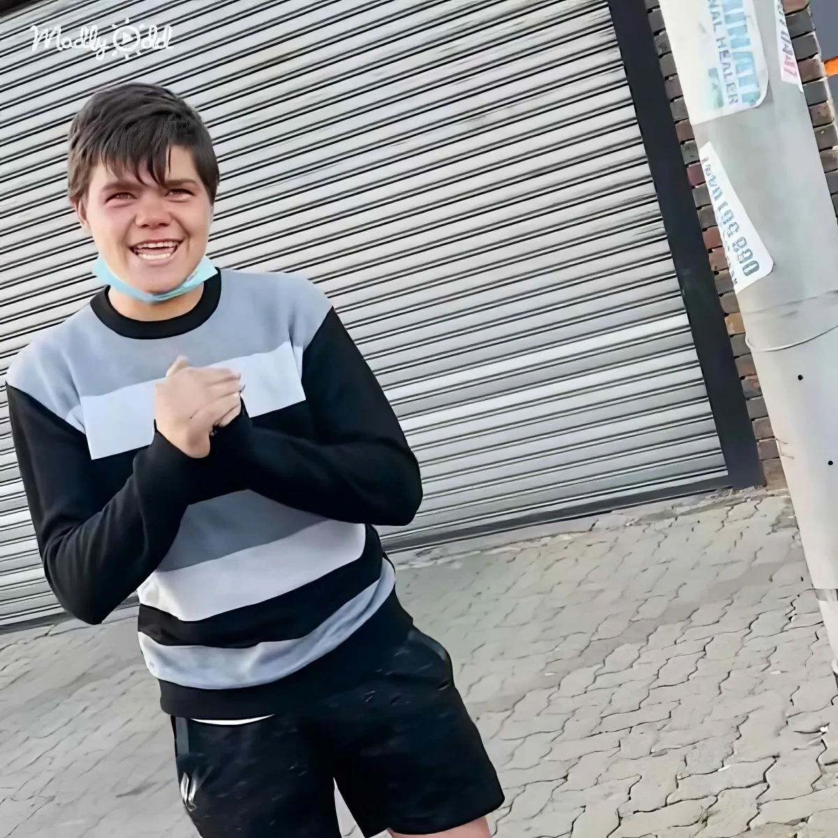A boy in a striped sweater and shorts smiles while holding his hands together in front of him.