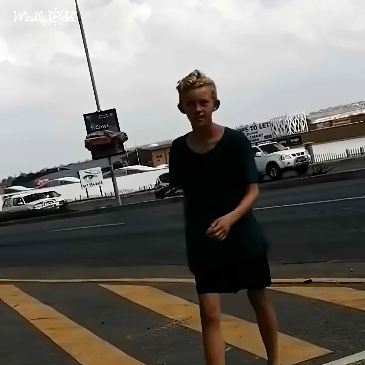 A person in a black dress stands on a crosswalk with a cloudy sky and parked cars in the background.