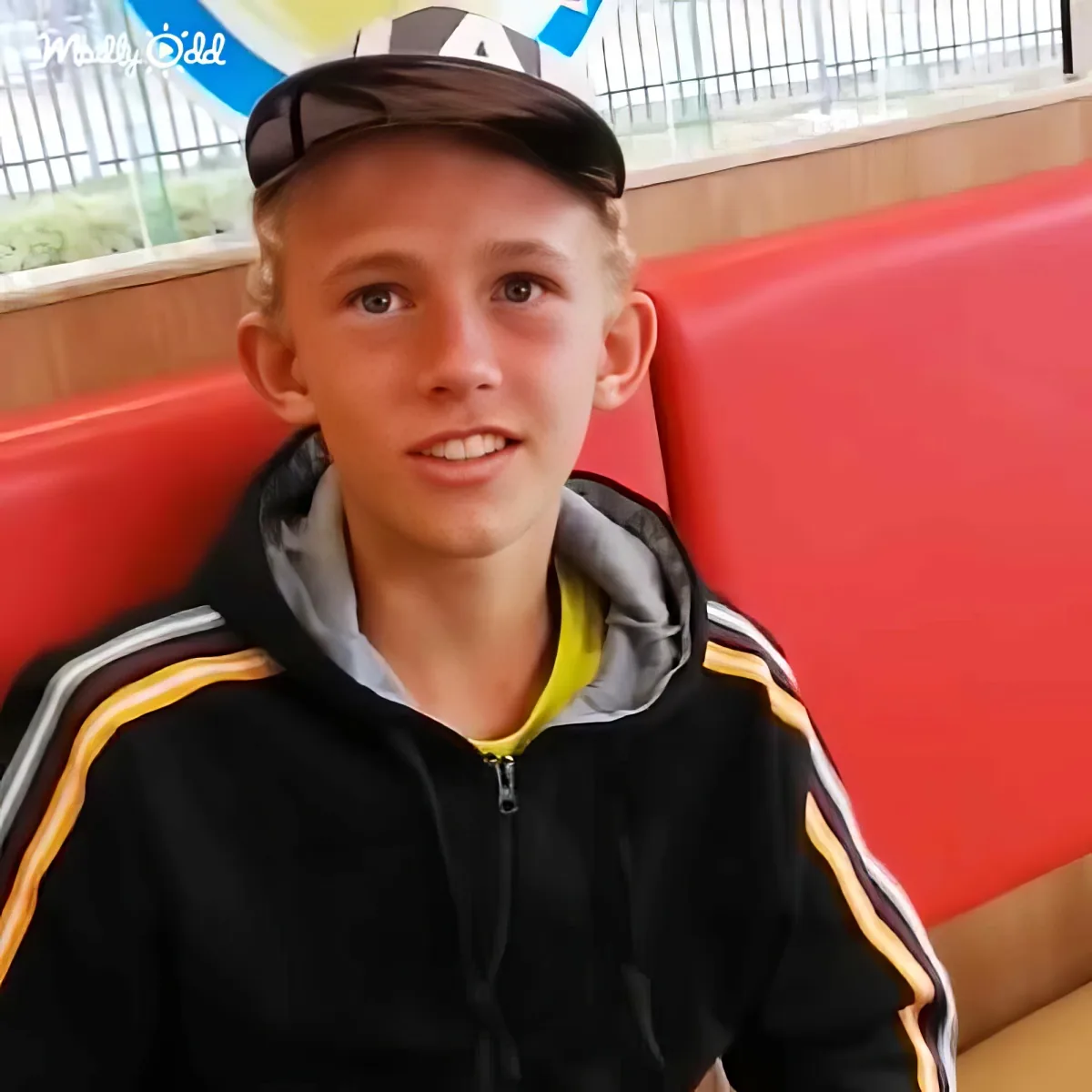 A young person wearing a black jacket with yellow and white stripes sits on a red bench, wearing a cap.