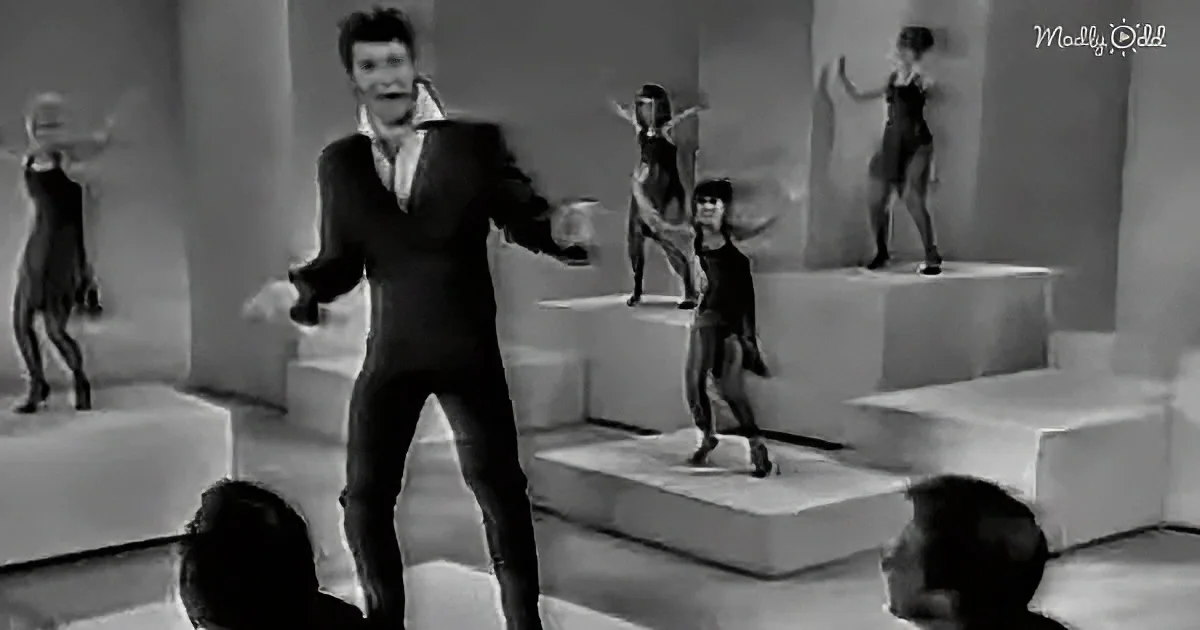 A man in a black suit dances energetically on a stage with dancers on white platforms behind him.