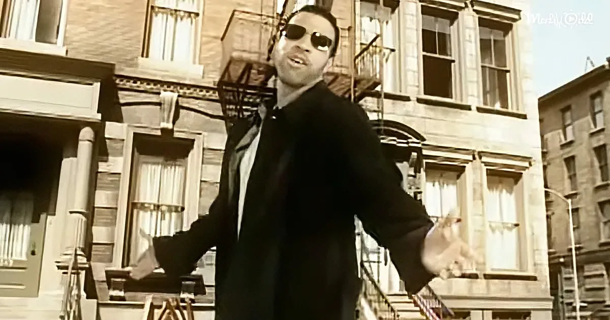 A man in a black coat and sunglasses stands in front of a row of buildings, arms outstretched.