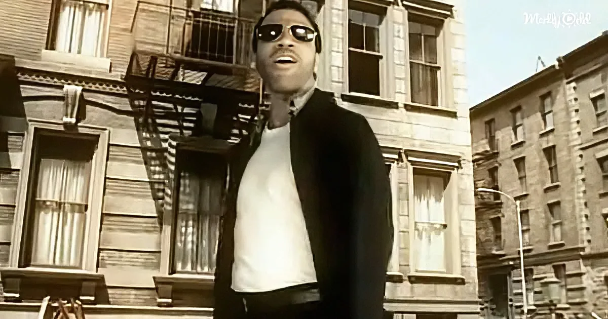 A man in sunglasses and a black jacket stands in front of a building with fire escapes.