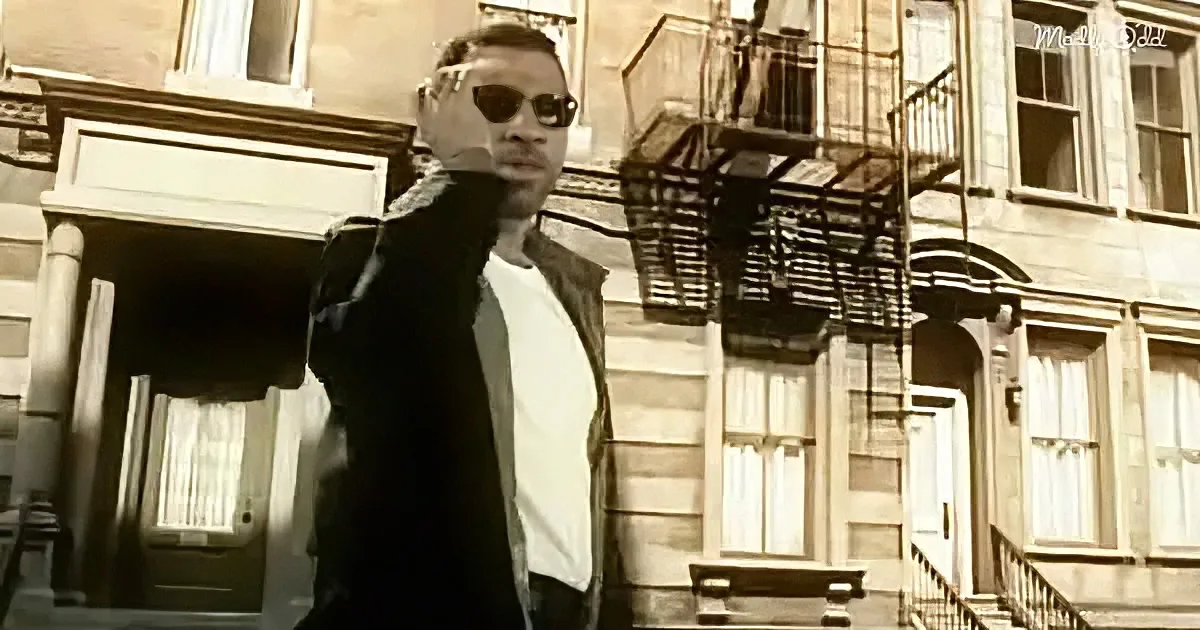 A man in sunglasses and a black jacket stands in front of a stone building with fire escapes.