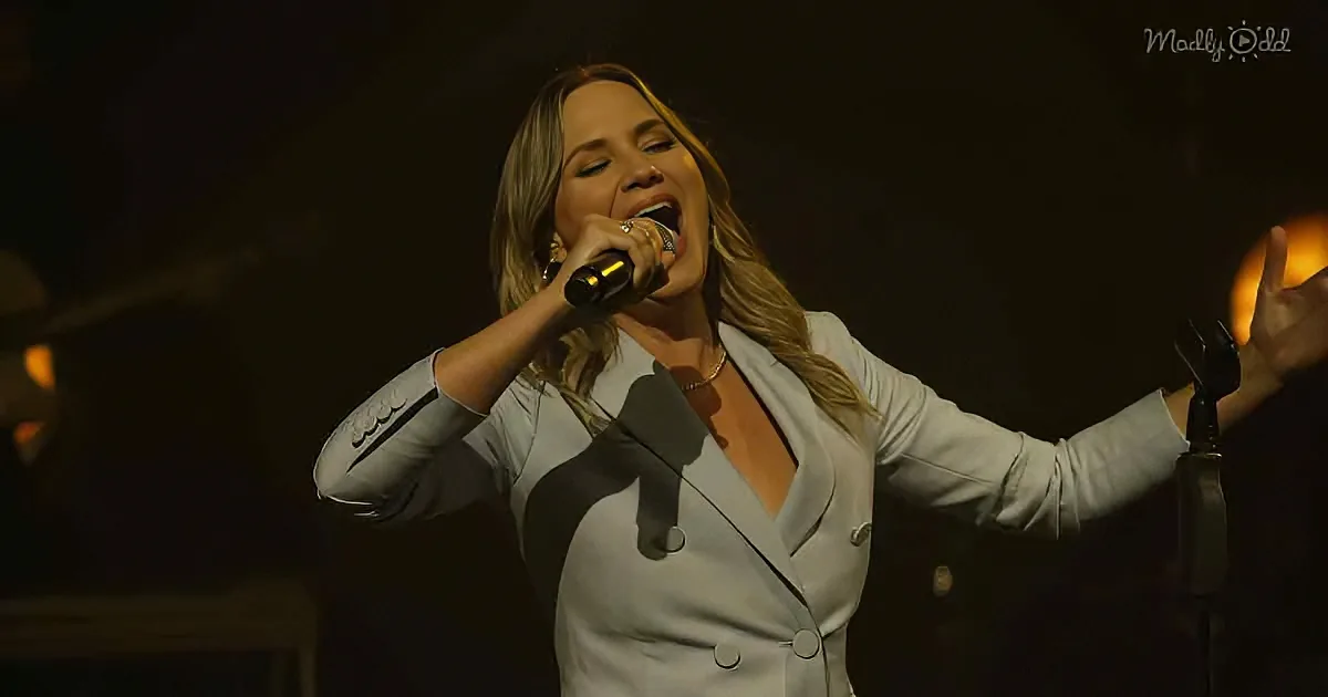 A woman in a white blazer sings passionately into a microphone on stage.