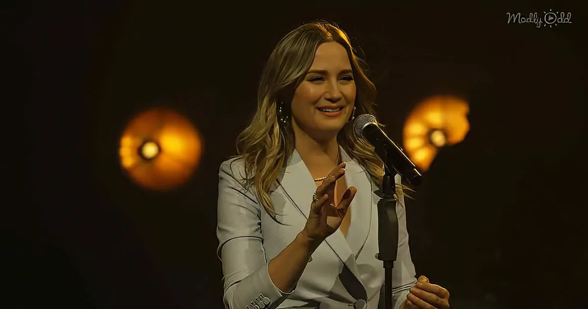 A woman in a white blazer smiles while gesturing near a microphone, with warm lights in the background.