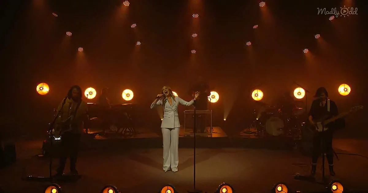 A woman in white sings on stage with a band, surrounded by warm lighting and smoke.