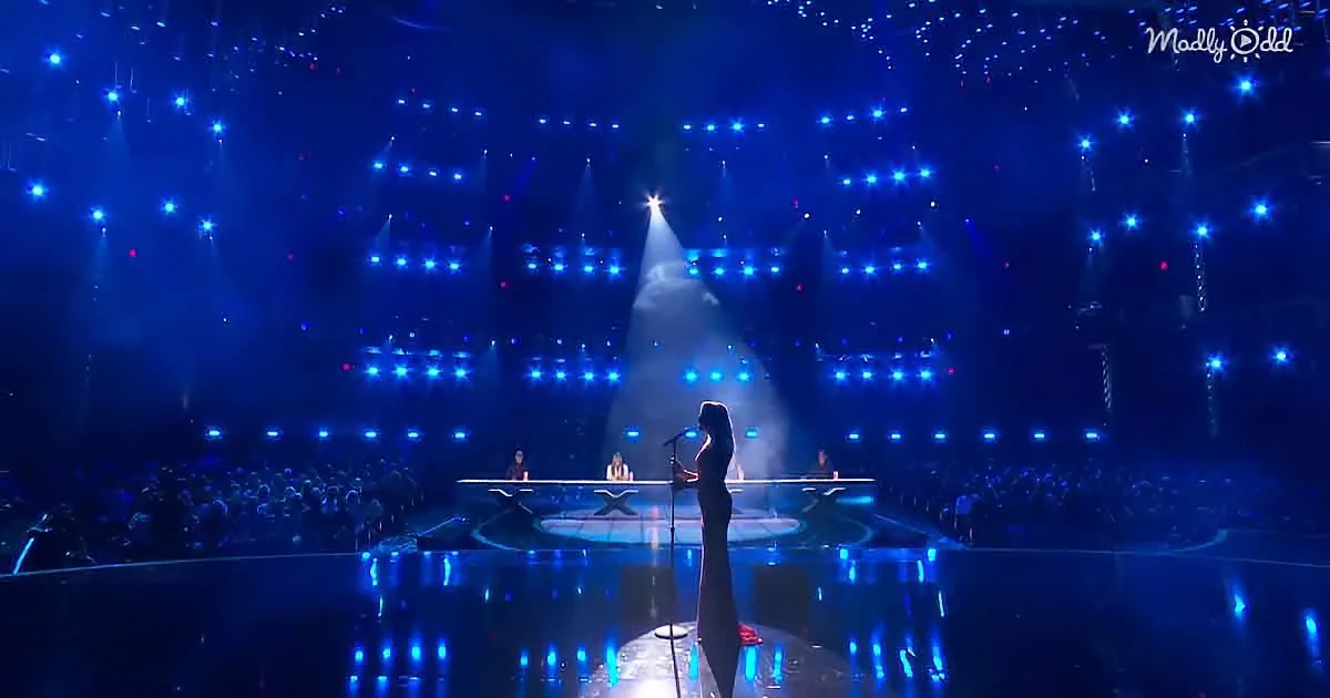 A silhouette of a woman stands on a stage with blue lights and a spotlight, facing a panel of judges.