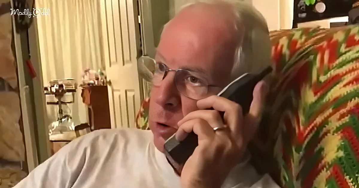 A man in a white shirt sits on a floral-patterned couch, holding a phone to his ear, appearing to be engaged in a...