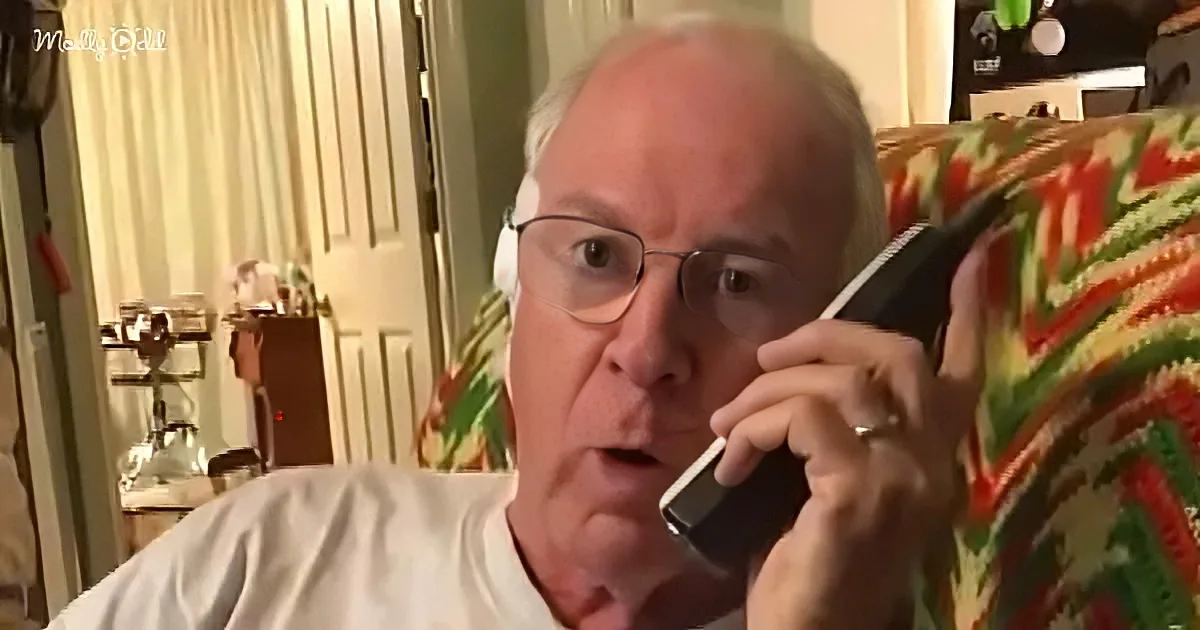 An older man with glasses and white hair is talking on a black phone while sitting on a colorful couch.