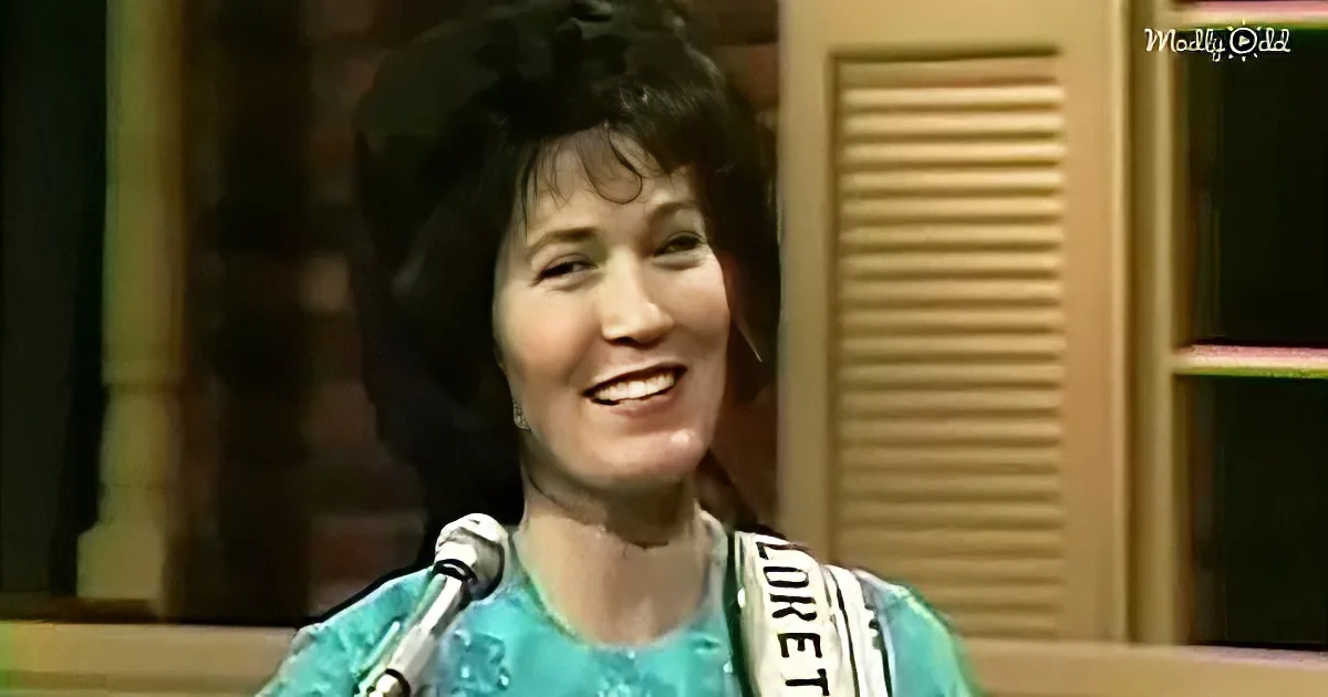 A smiling woman in a teal dress with a sash, standing in front of a microphone.