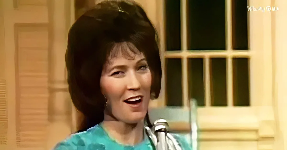 A woman with short, dark hair sings into a microphone, wearing a teal top, with a window and door in the background.