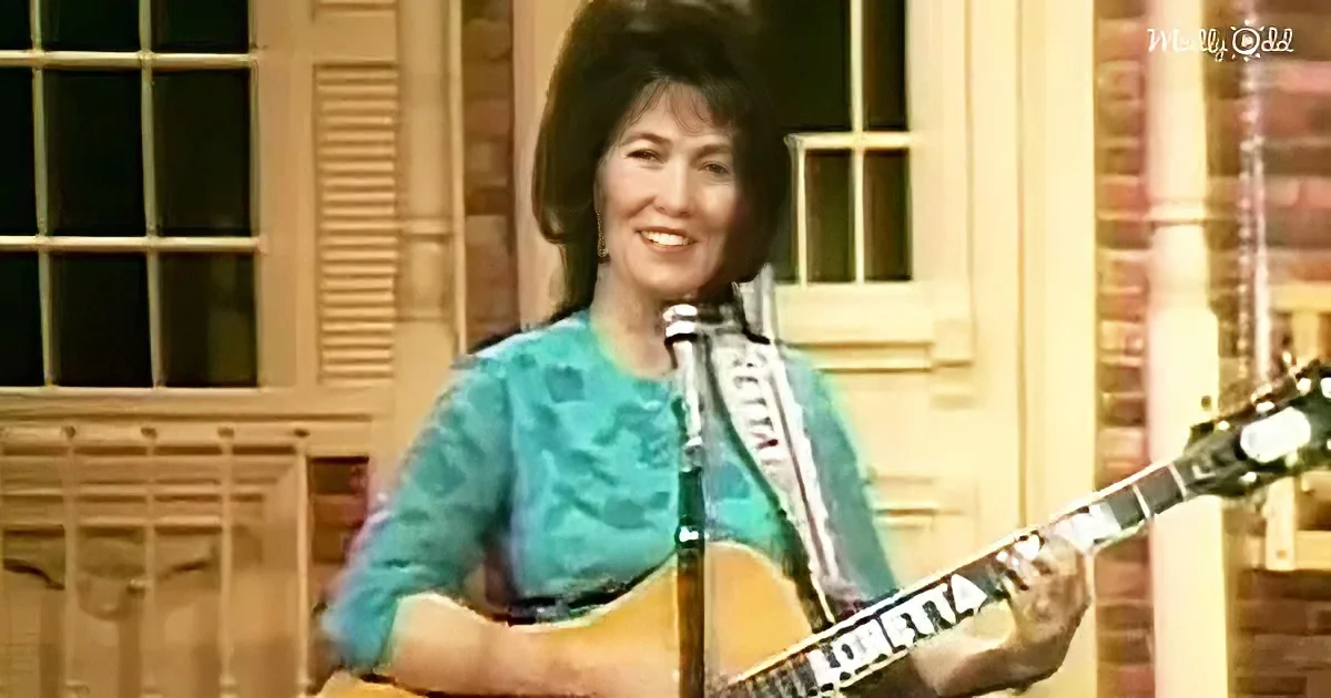 A woman in a teal dress plays a guitar on a stage with a microphone and a brick building in the background.