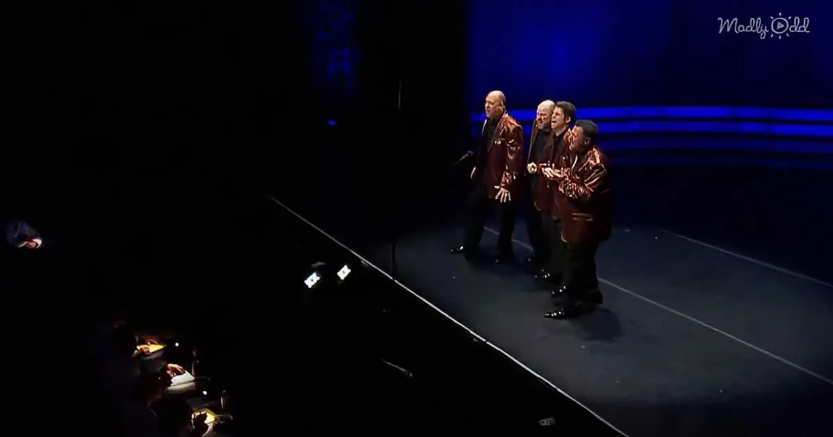 Four men in brown jackets sing on stage under blue lighting.