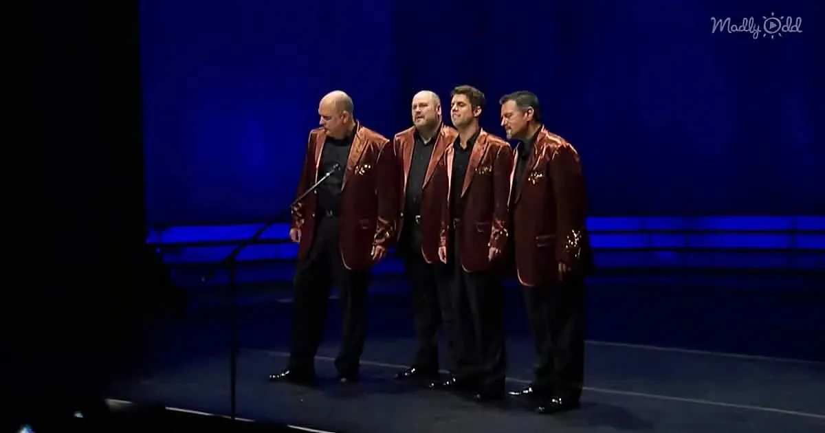 Four men in shiny red jackets stand on stage, singing into a microphone.