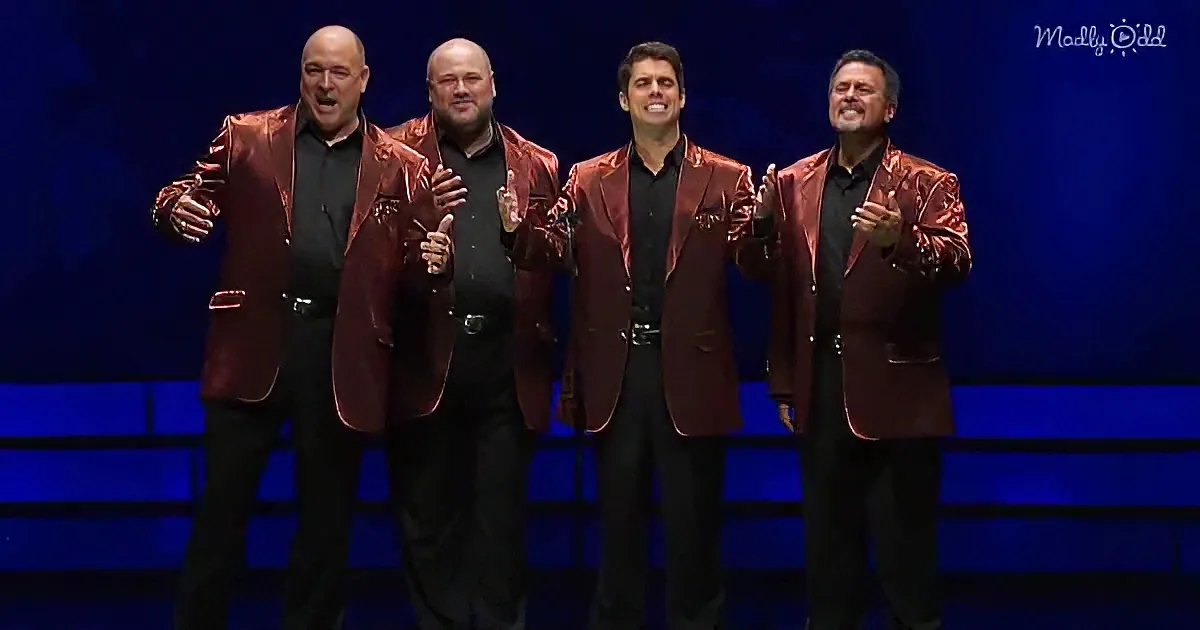 Four men in shiny red jackets and black shirts stand on stage, smiling and gesturing.