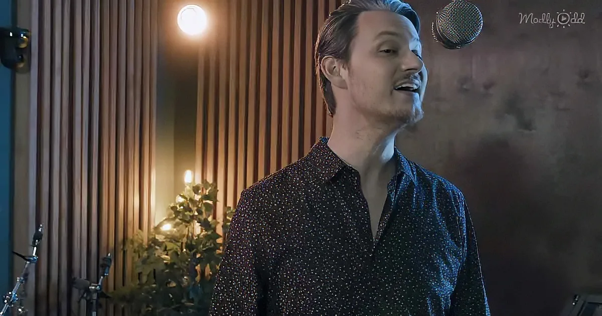 A man in a patterned shirt sings into a microphone in a warmly lit room with vertical wood panels.