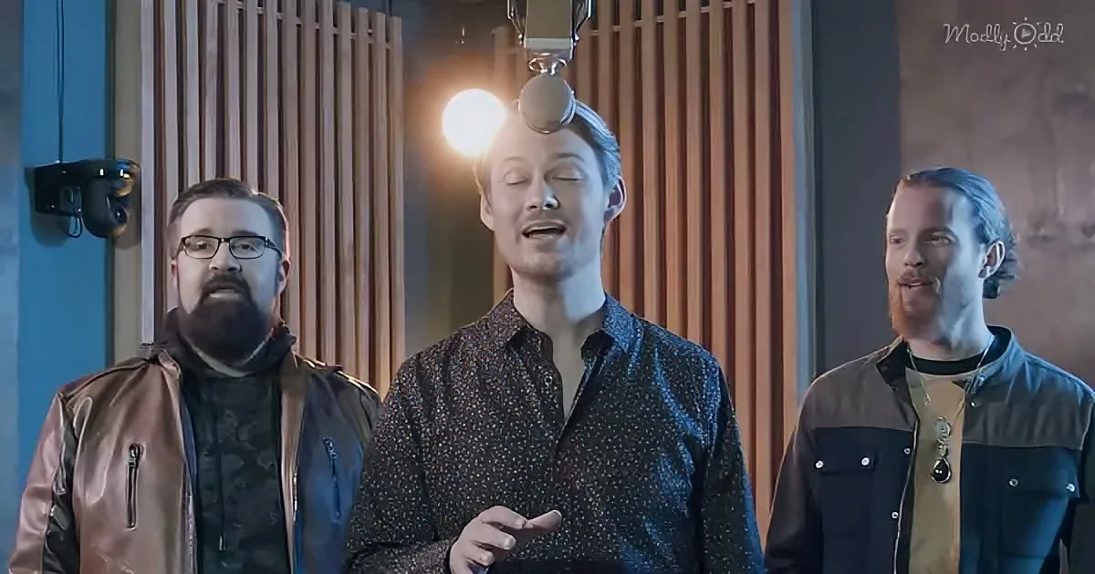 A man in a patterned shirt sings into a microphone, flanked by two others in a recording studio setting.