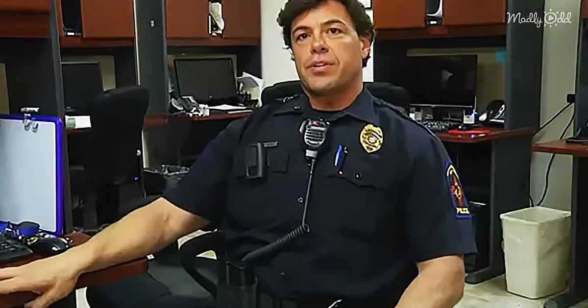 A police officer in uniform sits at a desk in an office, holding a device.