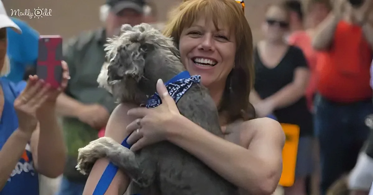 A woman smiles widely, holding a small, scruffy dog, surrounded by a crowd of people.