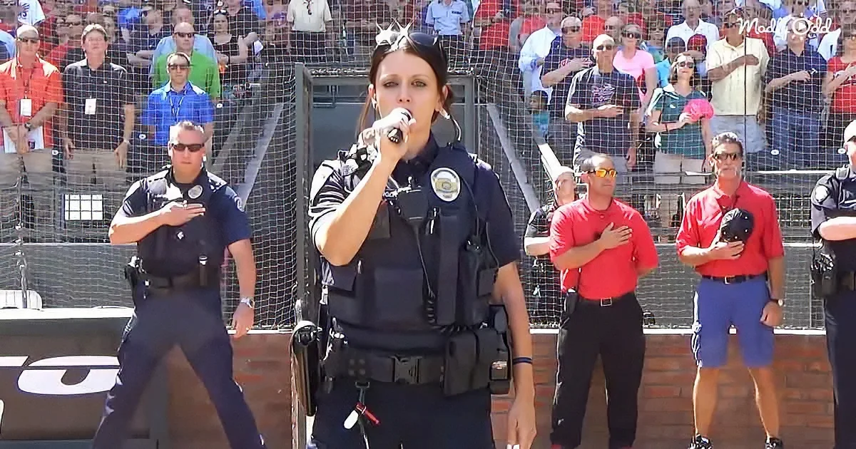 A woman in police uniform sings into a microphone while a crowd and security personnel stand behind her.