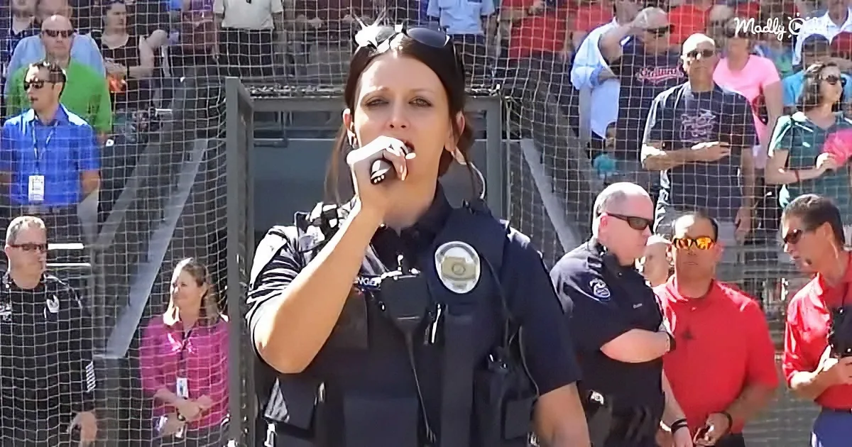 A woman in a police uniform sings into a microphone at a public event, surrounded by spectators.