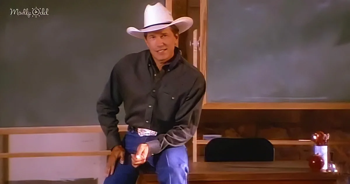 A man in a cowboy hat and blue jeans sits on a desk, smiling.