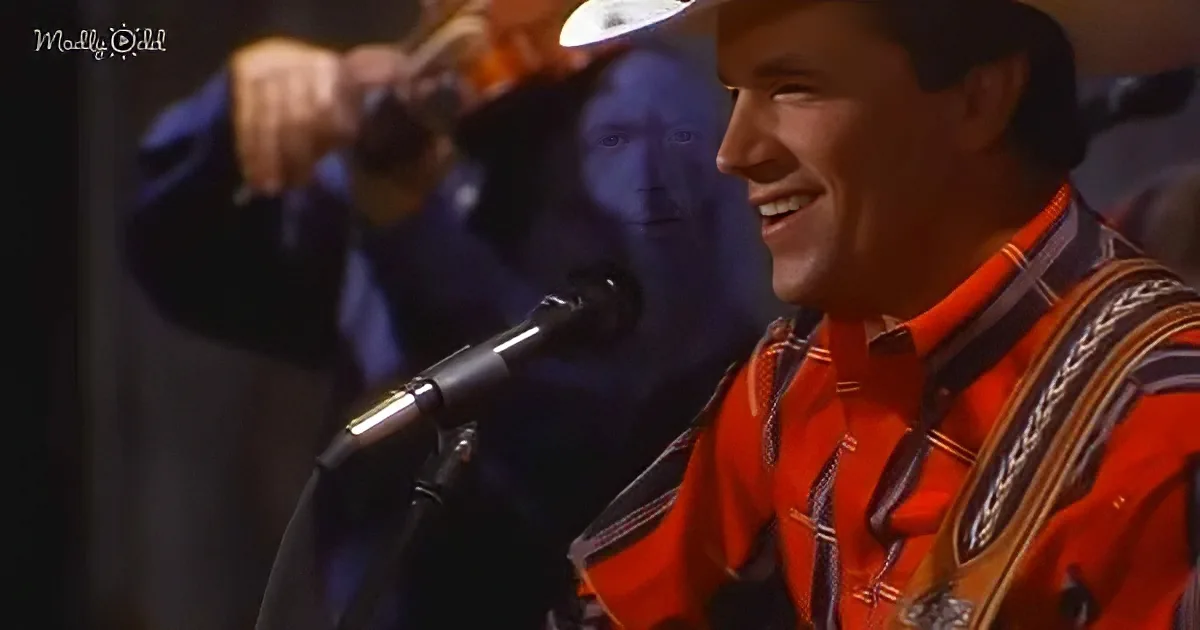 A man in a red shirt and hat smiles while playing a guitar in front of a microphone.