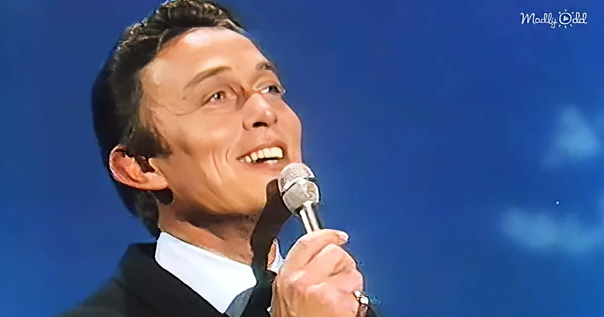 A man in a suit holds a microphone, smiling and looking upwards against a blue sky.
