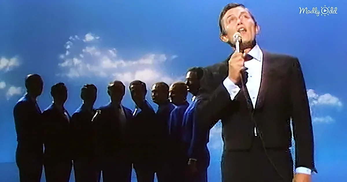 A man in a suit sings into a microphone, silhouetted against a blue sky with clouds.