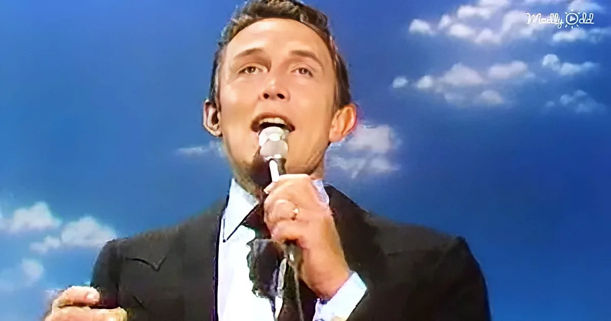 A man in a suit sings into a microphone against a blue sky backdrop.