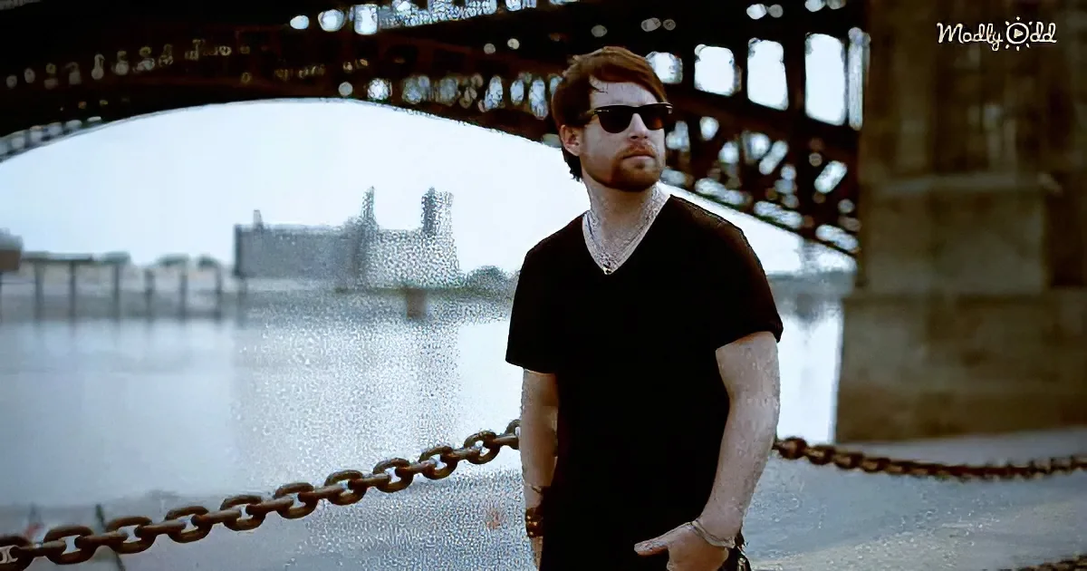 A man in a black shirt and sunglasses stands by a river with a bridge in the background.
