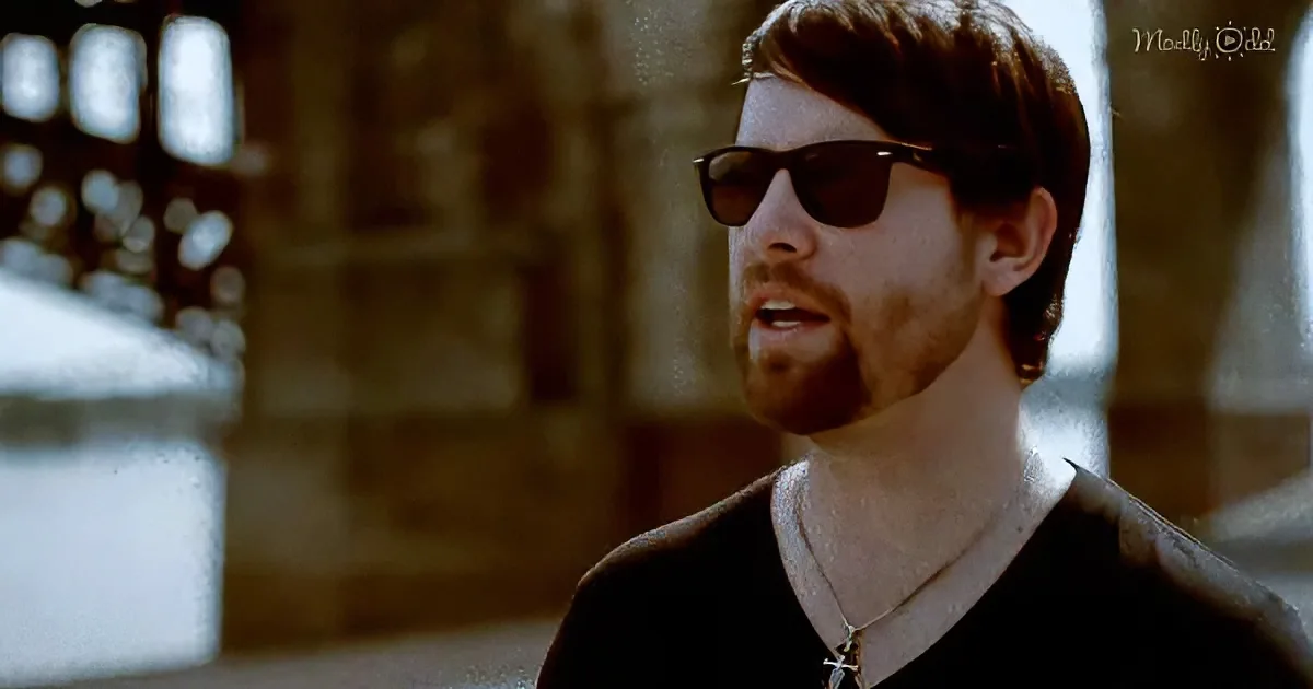A man with sunglasses and a beard, wearing a black shirt and a necklace, stands in a dimly lit, possibly historical setting.
