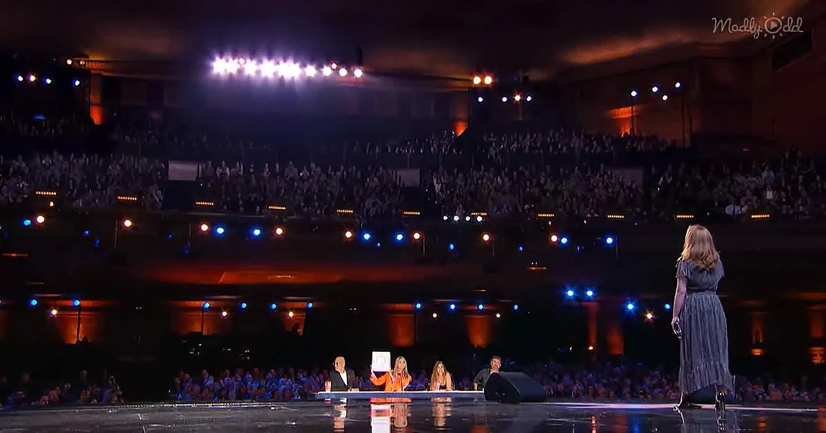 A woman in a blue dress stands on stage, facing judges and a large audience in a dimly lit arena.