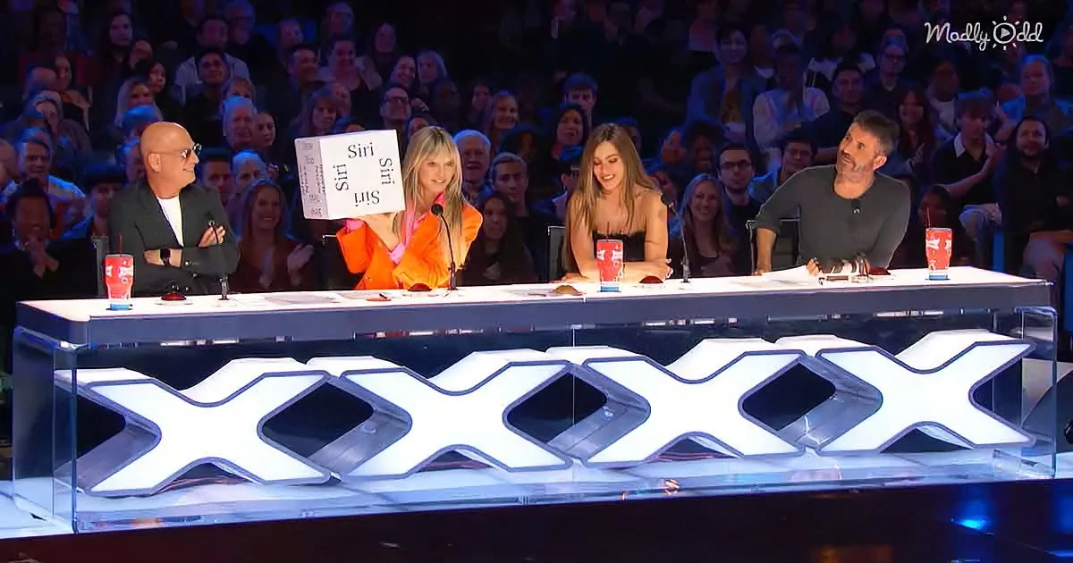 A woman holds up a sign reading "Siri" while seated at a panel table with judges, in front of a large audience.