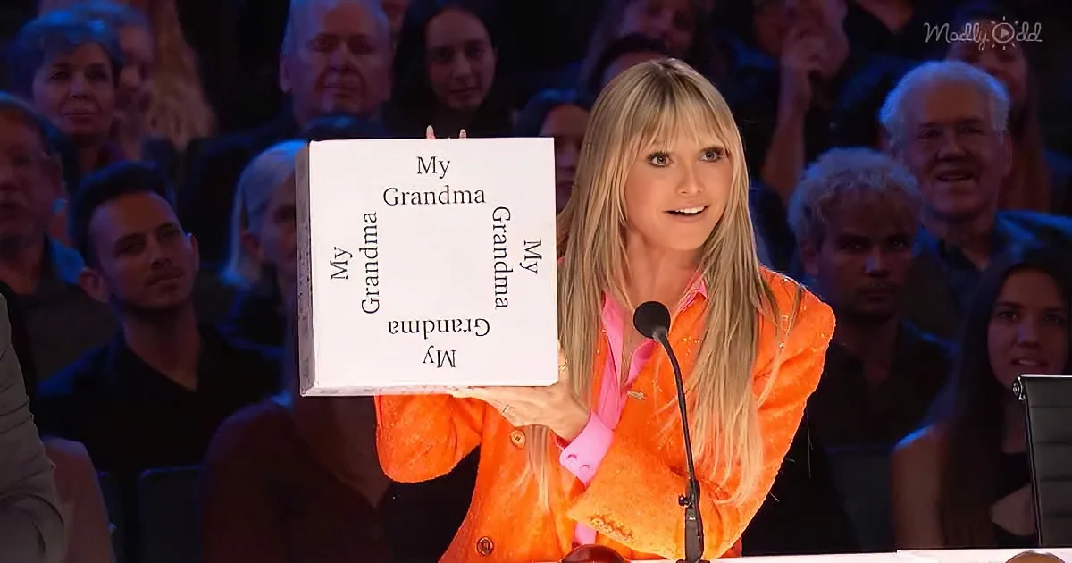 A woman in an orange outfit holds a sign saying "My Grandma" in front of a seated audience.