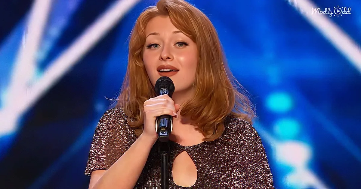 A woman with red hair holds a microphone, wearing a sparkly dress, on a stage with blue lights.