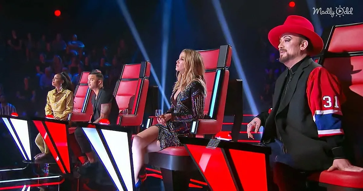 Boy George in red hat stands beside seated judges on red chairs in a studio setting.