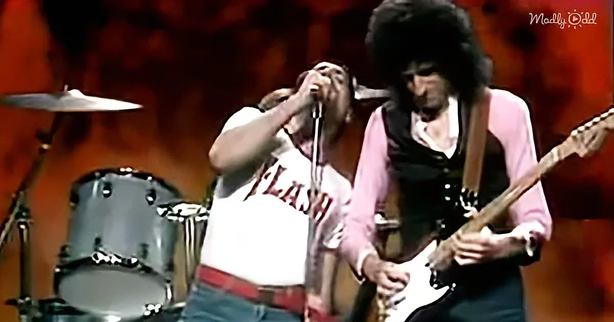A man in a white shirt with "FLASH" sings into a microphone while another man plays guitar on stage.
