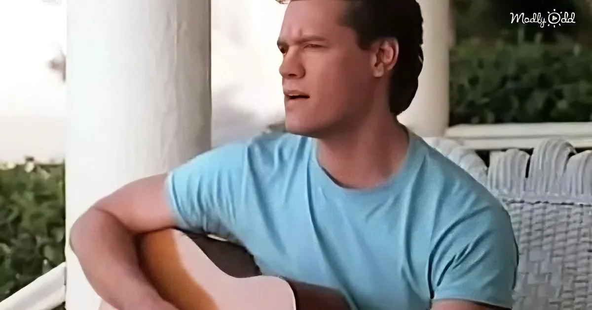 A man in a light blue shirt sits on a porch, holding a guitar, with a white railing and greenery in the background.