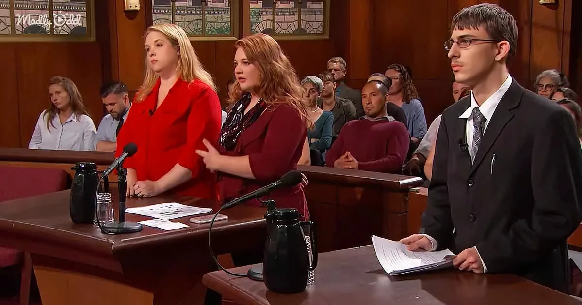 A man in a suit stands at a desk with papers, while two women in red stand behind him, and an audience sits in the...