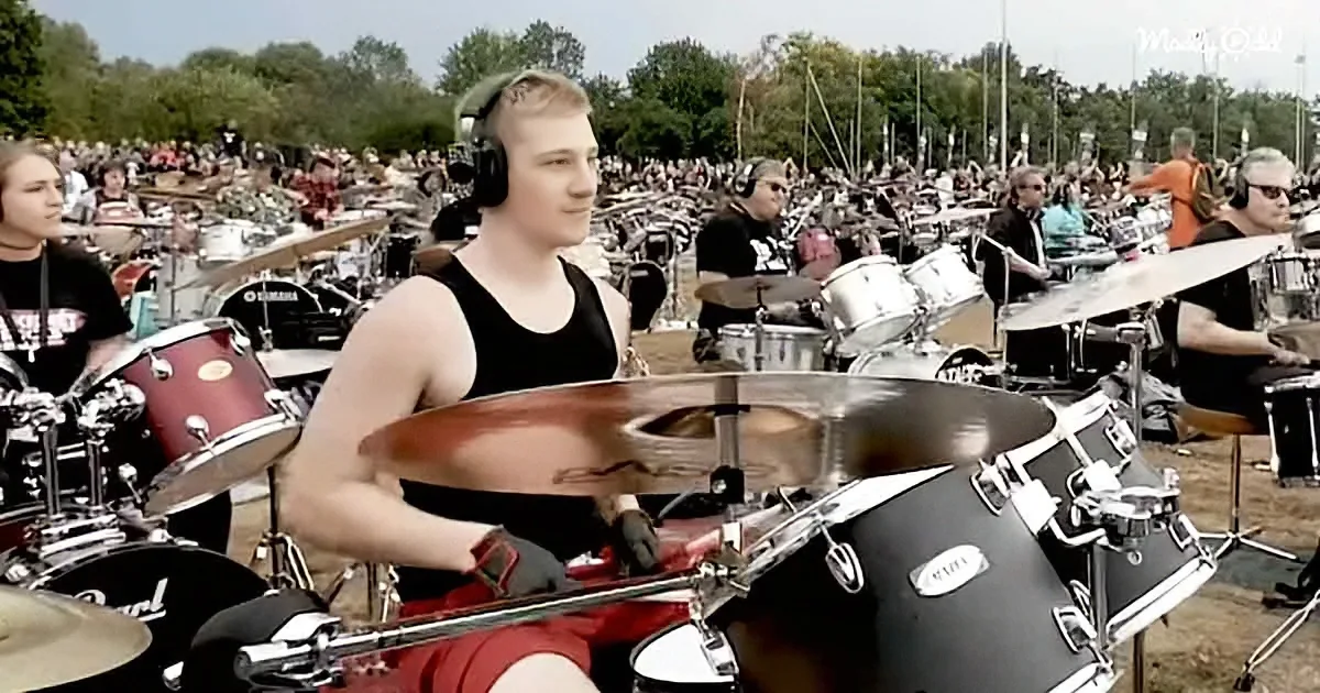 A drummer in a black tank top plays a drum set in front of a large crowd of other drummers at an outdoor event.