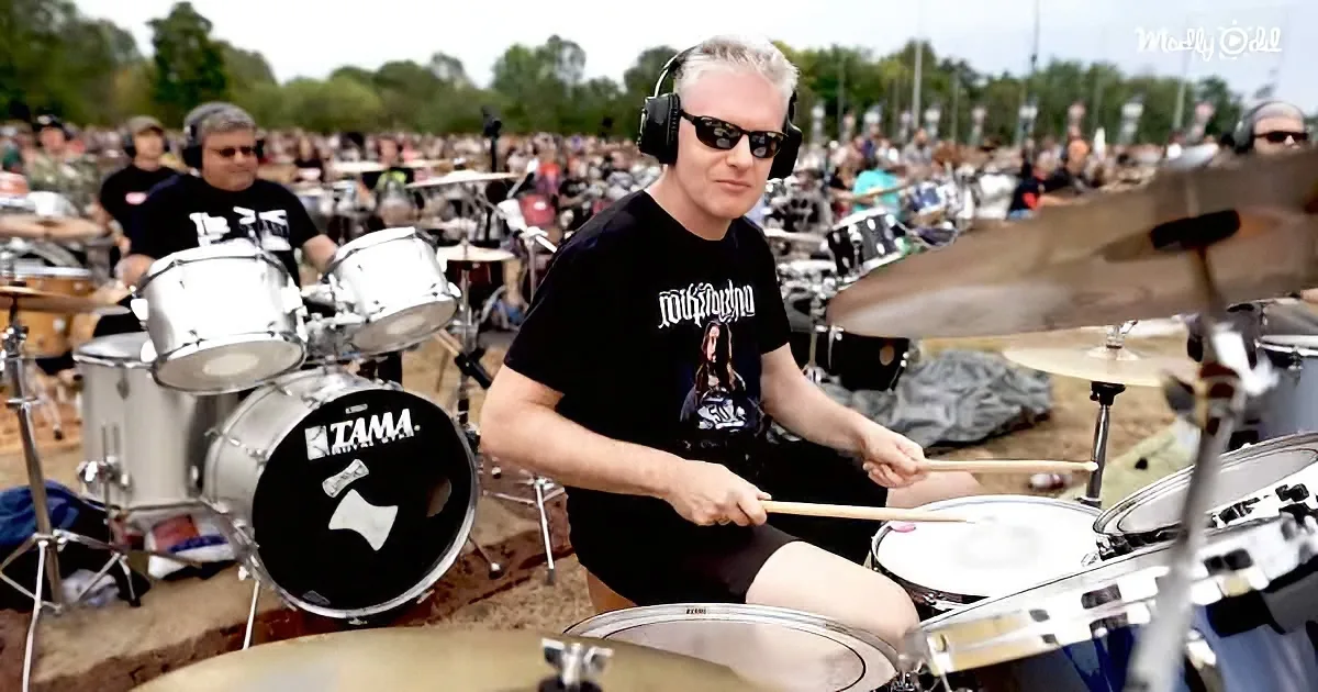 A man wearing headphones and sunglasses plays drums in front of a large crowd at an outdoor event.