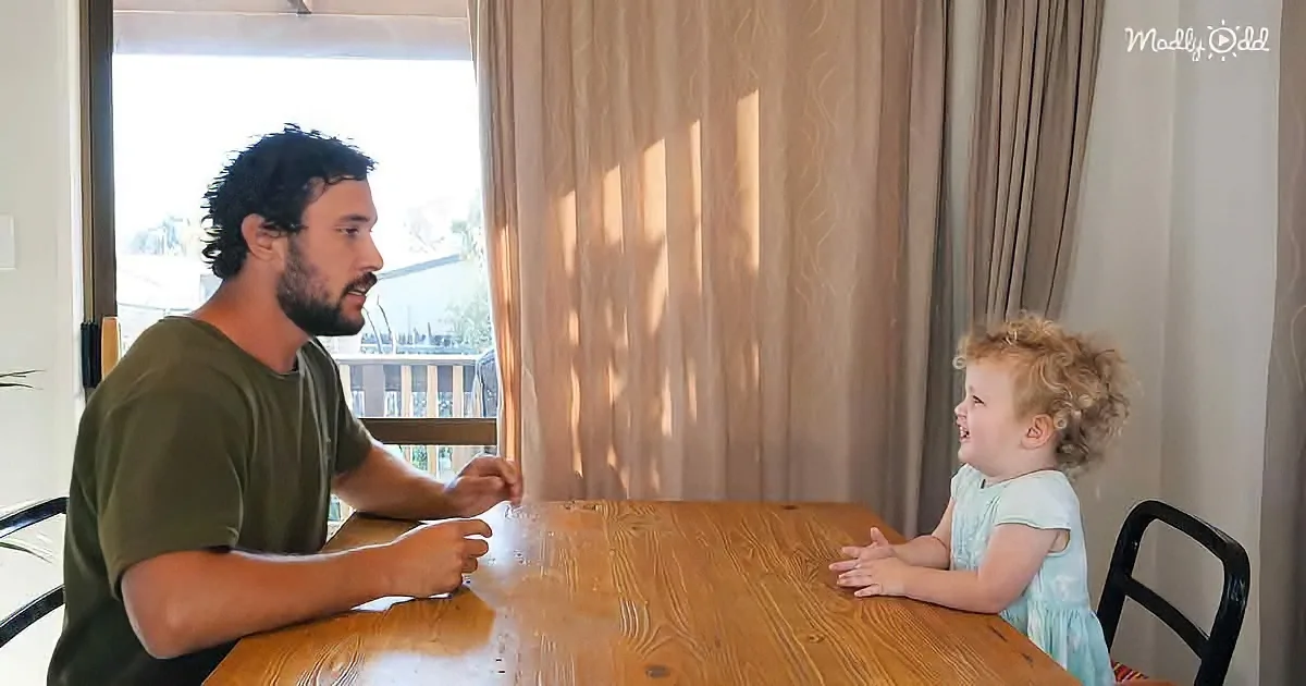 A man and a child sit at a wooden table, facing each other, in a room with curtains and a window.