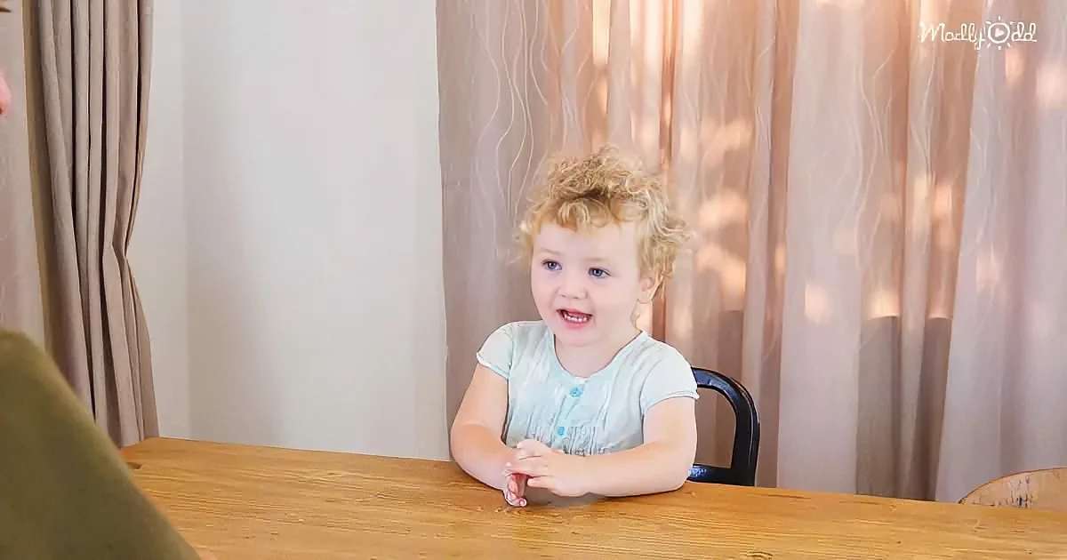 A smiling toddler with curly hair sits at a wooden table, hands clasped, in front of light curtains.
