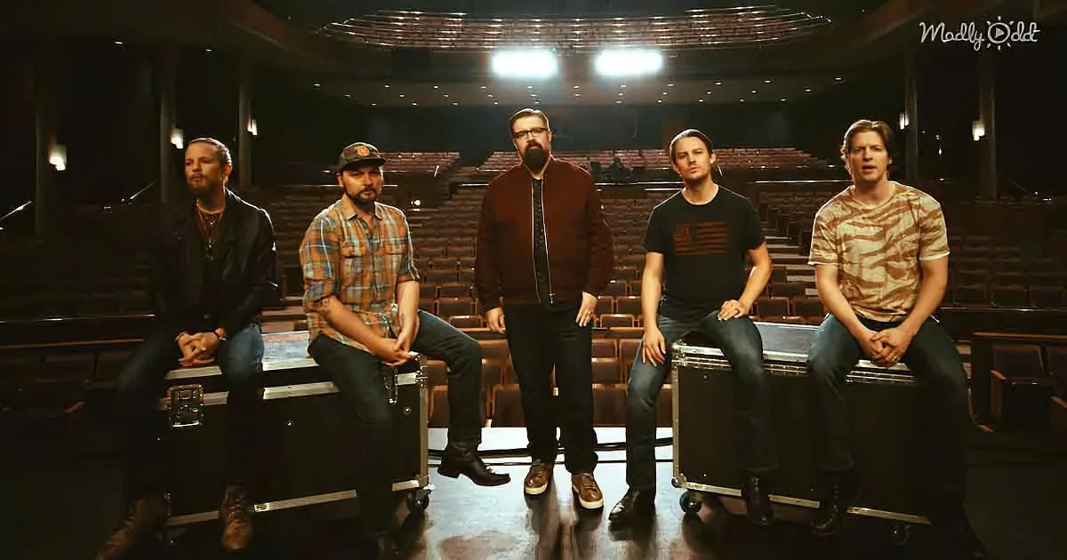 Five men in casual attire sit on stage cases in an empty auditorium, facing forward.