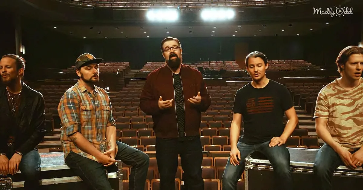 A man in a maroon jacket stands center stage, gesturing, surrounded by seated men in casual attire in an empty auditorium.