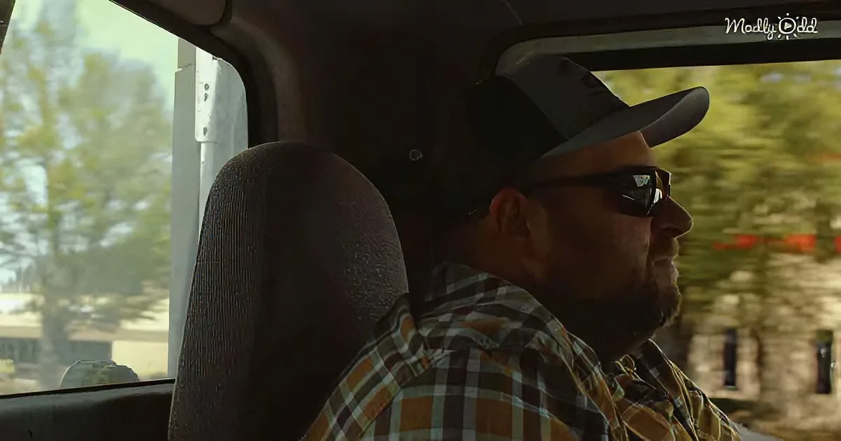 A man wearing a hat and sunglasses sits in a vehicle, looking out the window.